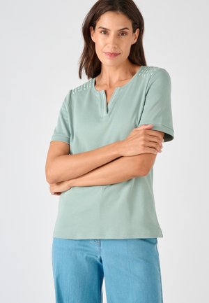 Vrouw staat met gekruiste armen, draagt een lichtgroen kort mouwtopje met schouderdetails en lichtblauwe jeans tegen een effen achtergrond.