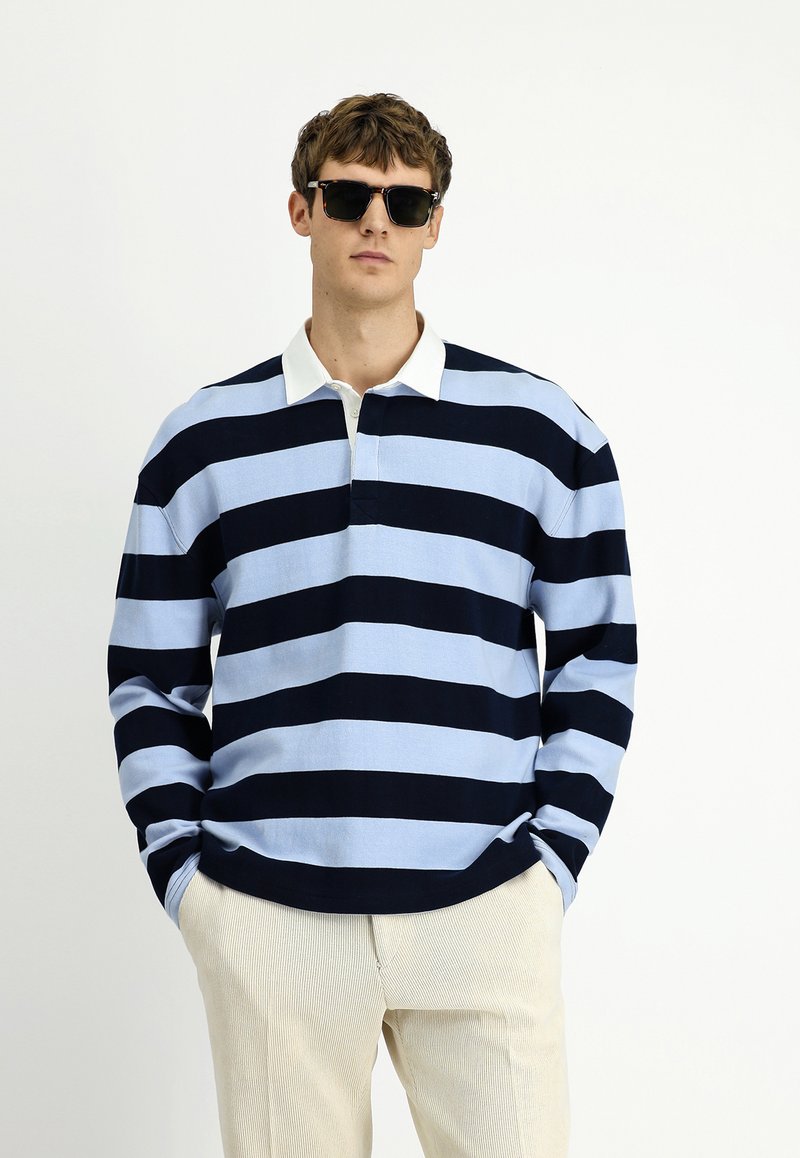 Polo a strisce blu navy e celeste con maniche lunghe e colletto bianco, dotato di patta con bottoni. Indossato con pantaloni a strisce beige.