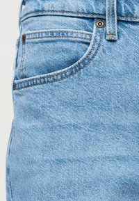 Κοντινή λήψη ανοιχτό μπλε τζιν παντελονιού που δείχνει την μπροστινή τσέπη, τα νύχια, την θηλιά της ζώνης και τις πορτοκαλί ραφές πάνω στην υφή του υφάσματος.