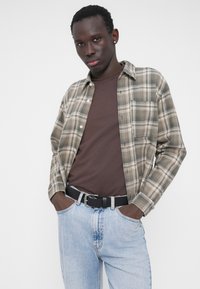 Koszula w kratę w beżowych i brązowych odcieniach, noszona na brązowym t-shircie. Jasnoniebieskie dżinsy z czarnym paskiem. Widoczne szczegóły tekstury materiału.