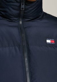 Marinblå pufferjacka med hög krage, full dragkedja och logotypmärke. Slät yta med kviltad yta och subtilt rutmönster.