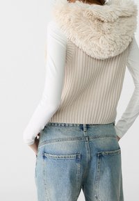 Mujer con un chaleco de punto beige sin mangas y con capucha esponjosa, sobre una camiseta blanca de manga larga y jeans de mezclilla azul claro, vista desde atrás.