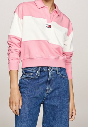 Persoon draagt een roze en wit kleurblok langärmelig poloshirt met Tommy Jeans-logo en blauwe high-waisted spijkerbroek.
