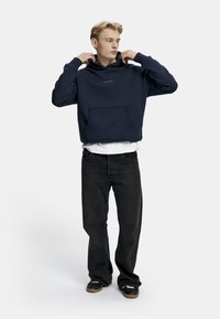 Mørkeblå hettegenser med frontlomme, løs passform og stor hette, båret over en hvit t-skjorte; parret med svarte jeans og svarte sko.