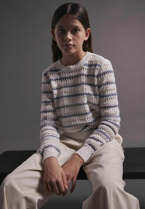 Pleteni pulover u krem boji s plavim i zelenim prugama, s teksturiranim uzorkom i rebrastim manžetama, uparen s svijetlim hlačama.