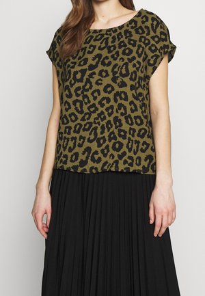 Vrouw draagt een korte mouw olijfgroen luipaardprinttop gecombineerd met een zwarte geplooide rok, staand tegen een effen achtergrond.