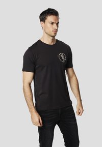 Black short-sleeve t-shirt made of cotton with a round neck. Features a graphic design on the left chest area. Worn with dark jeans.