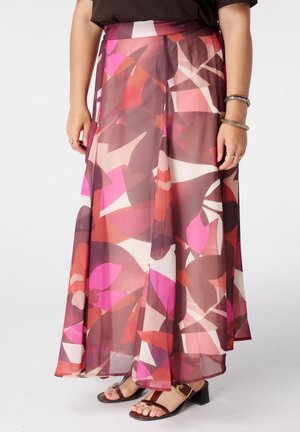 Femme portant une longue jupe fluide avec des formes abstraites roses, rouges et brunes, associée à des sandales à talons noires et des bracelets aux poignets.