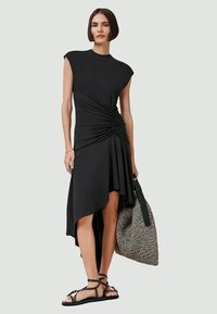 Vestido preto com mangas em cap sleeve, bainha assimétrica e detalhe franzido na lateral. Apresenta uma textura suave. Combinado com uma mala de mão estampada.