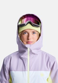 Jasnofioletowa kurtka snowboardowa z kapturem, biała dolna część oraz żółte akcenty. Towarzyszy jej żółta czapka i lustrzane gogle narciarskie.