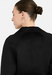 Abrigo de lana negro con cuello, que presenta una textura suave y hombros estructurados. Se pueden ver pendientes largos de cadena plateada.