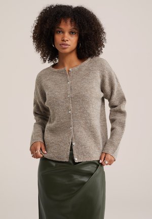 Femme aux cheveux bouclés portant un pull beige boutonné et une jupe en cuir vert foncé, debout devant un fond neutre.