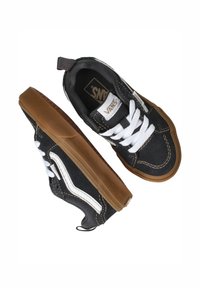 Zapatillas de lona negras con cordones blancos, franja lateral blanca en contraste, suela de goma marrón y detalles de costura. La etiqueta en la lengüeta dice "VANS."