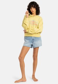 Yellow cotton hoodie with a graphic design in orange and teal, paired with light blue denim shorts with a frayed hem.