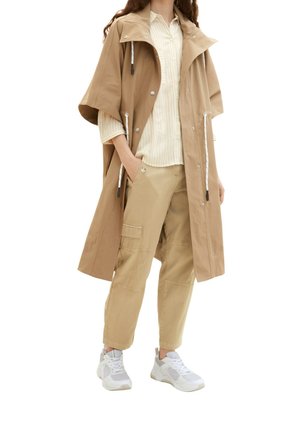 Abrigo impermeable beige con capucha, mangas cortas, detalles en plata, bolsillos laterales, superpuesto sobre una camisa de rayas ligera y pantalones caqui, combinado con zapatillas blancas.