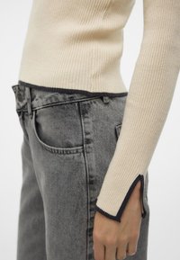 Maglione beige a coste con bordi neri sui polsi, abbinato a jeans grigi a vita alta con leggeri sbiadimenti e tasche laterali.