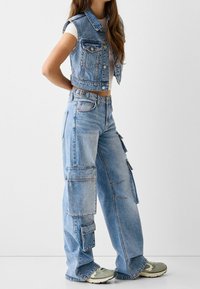 Jeansová vesta a cargo džíny ve světlé modré. Vesta má přední kapsy a zkrácený střih; džíny mají více bočních kapes a volný střih.