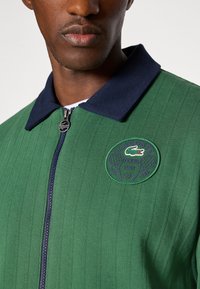 Casaco riscado verde com colarinho azul-marinho, fecho éclair prateado e um emblema redondo do Lacoste Tennis Club, apresentando raquetes de ténis e o logótipo.