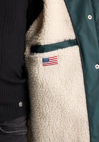 Green quilted vest with a cream sherpa interior, featuring a small American flag patch and a contrasting fabric pocket. Black ribbed shirt visible.
