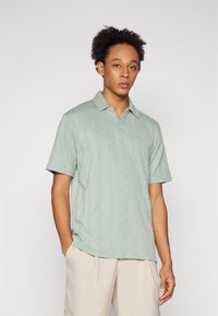Polo de manga curta verde claro com um padrão de diamante texturizado e colar clássico. Usado com calças beige claras.