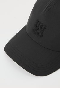 Melna auduma cepure ar nedaudz teksturētu virsmu. Priekšpusē ir uzmontēts melns "HUGO" logo un izšuvumi.