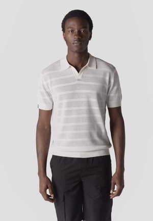 Fiatal férfi, fehér, rövid ujjú, vízszintes csíkos galléros pólót és fekete cargo nadrágot visel, egyszerű háttér előtt áll.