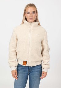 Giacca in pile color crema con colletto alto, chiusura con zip, polsini a coste e un'etichetta arancione sull'orlo; materiale morbido e testurizzato.