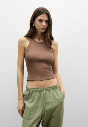 Jeune femme aux longs cheveux bruns portant un crop top marron sans manches et un pantalon vert ample, les mains dans les poches, sur fond uni.