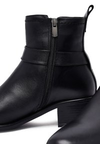 Bottes noires en cuir à tige mi-molle, avec un petit talon bloc, une fermeture éclair latérale et deux sangles horizontales autour de la partie supérieure. Texture lisse, design élégant.