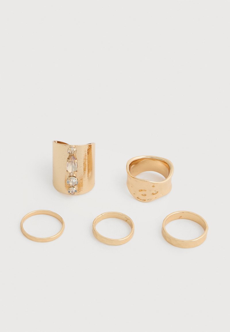 Cinq bagues en or de largeurs variées, dont une ornée de trois petites pierres précieuses transparentes, présentées sur un fond blanc.