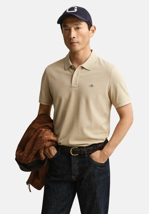 Homme portant un polo beige et une casquette bleue marine, tenant une veste marron dans la main gauche avec la main droite dans la poche de son jean.