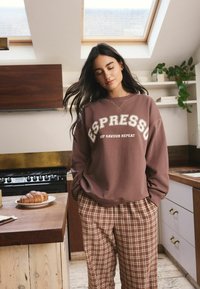 Brauner Sweatshirt mit weißem "ESPRESSO"-Text, gemusterte Karohose und ein Croissant auf einem Teller in einem modernen Küchenumfeld.