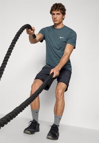 Idrettsmann i en tealfarget Nike t-skjorte og svarte shorts, som holder et tykt svart kamp tau. Grå sokker og svarte treningssko er også synlige.