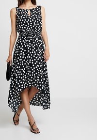 Robe à pois noirs et blancs avec un col en forme de trou, une taille élastique et un ourlet asymétrique, réalisée en tissu léger.