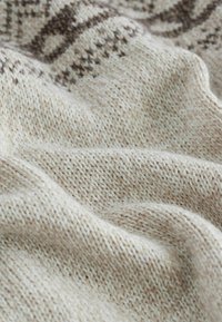Gros plan sur un tissu en maille beige doux avec un motif plus sombre subtil, montrant la texture et les plis à la lumière naturelle.