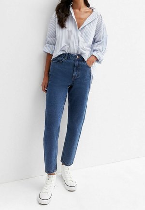 Femme portant une chemise à rayures bleues boutonnée, un jean bleu taille haute et des baskets blanches montantes, debout devant un fond blanc.