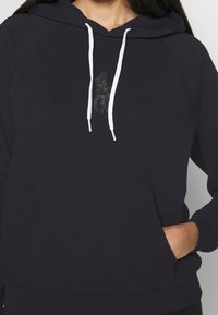 Marinblå hoodie med vita snörningar, framficka och en broderad ridsportsdesign på bröstet. Mjuk, texturerad tyg.