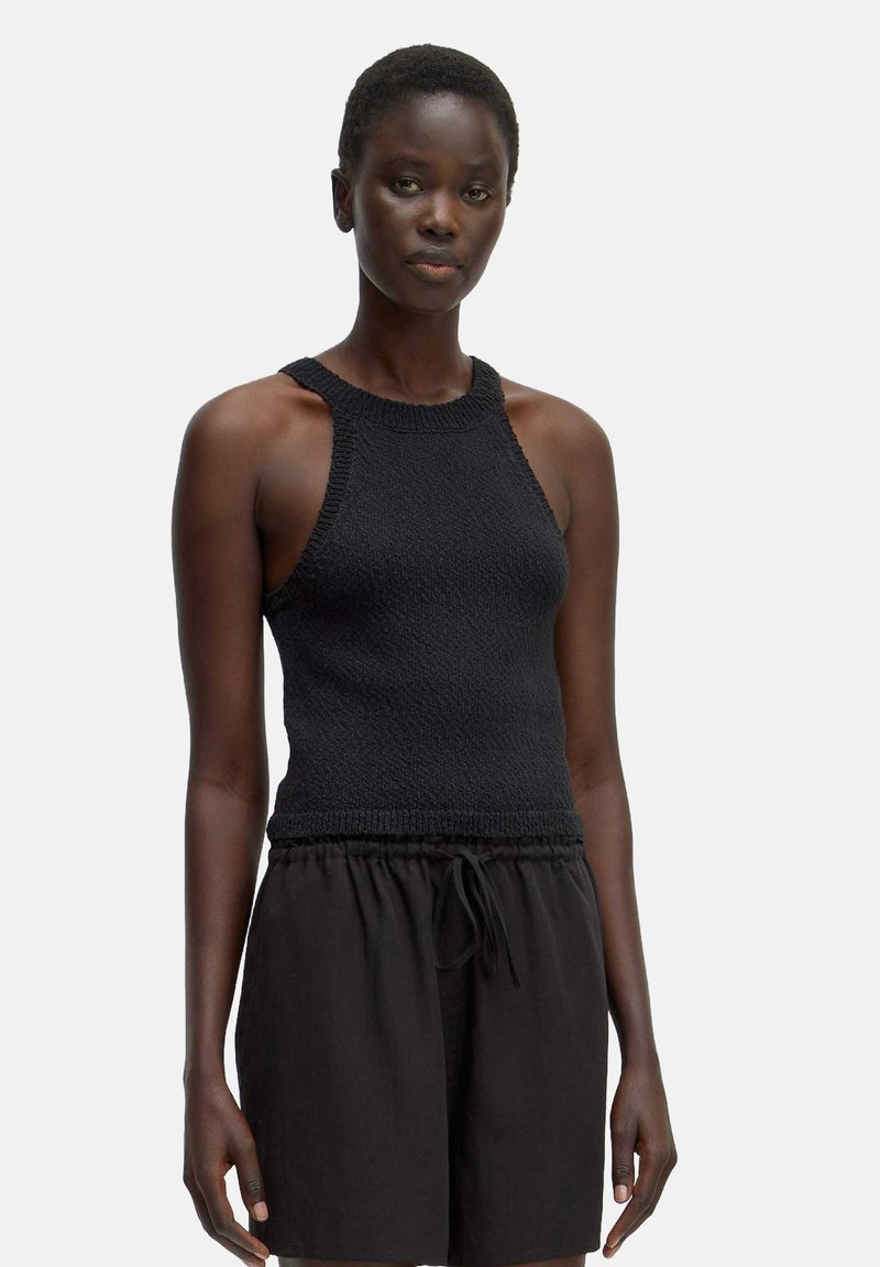 Schwarzes, gestricktes Tanktop mit hohem Ausschnitt, offenen Schultern und strukturiertem Stoff, kombiniert mit lockeren schwarzen Shorts.