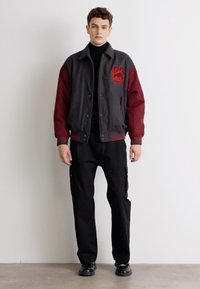 Gray and burgundy bomber jacket with logo on left chest, worn over a black turtleneck, paired with black cargo pants and shoes.