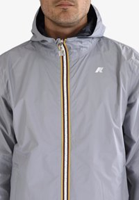 Homme portant une veste à capuche gris clair avec une fermeture éclair avant ornée de rayures rouges, jaunes et noires, et un petit logo blanc sur la poitrine.