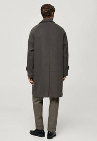 Gray wool overcoat with a high collar, raglan sleeves, buttoned cuffs, and a straight cut, worn over beige trousers and black shoes.