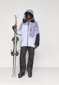 Άνδρας που φοράει γκρι και μαύρο μπουφάν σκι, μαύρο παντελόνι, μπότες σκι και γυαλιά σκι κρατώντας ένα ζευγάρι σκι όρθια μπροστά σε λευκό φόντο.