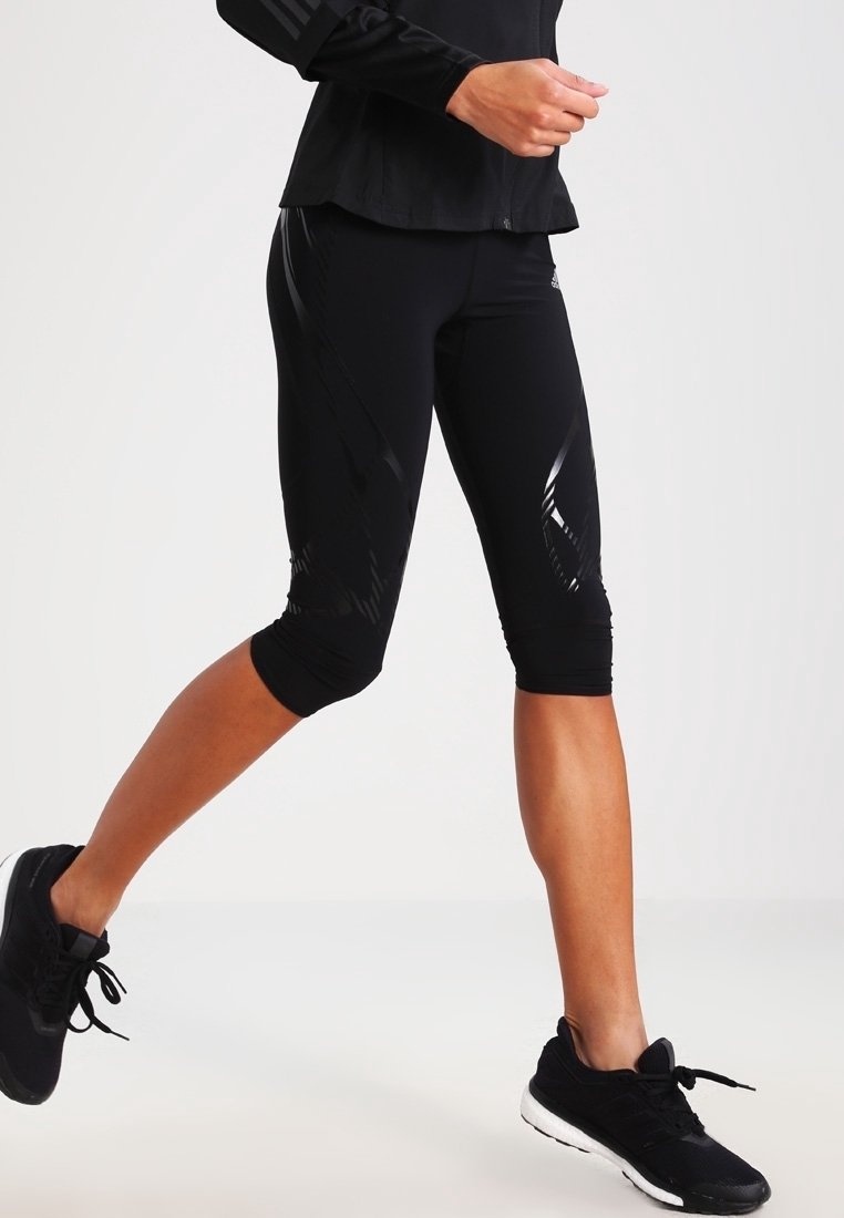 Persoon in zwarte sportcapri-leggings en zwarte hardloopschoenen, weergegeven in het midden van een stap tegen een effen achtergrond.
