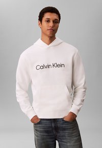 Jauns vīrietis ar īsiem cirtainiem matiem, valkājot baltu Calvin Klein kapuci un zilas džinsas, stāv ar rokām kabatās pelēka fona priekšā.