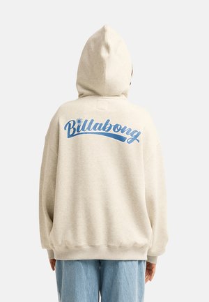 Osoba nosząca jasno beżową bluzę z kapturem z niebieskim logo "Billabong" na plecach, w zestawieniu z jasnoniebieskimi jeansami.