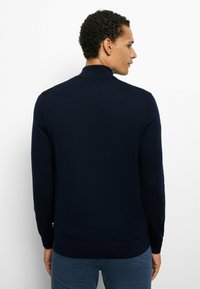 Dunkelblauer langärmeliger Polopullover mit geripptem Muster, ausgestattet mit einem kleinen Kragen und einem taillierten Saum. Einfaches Design, keine sichtbaren Logos oder Muster.