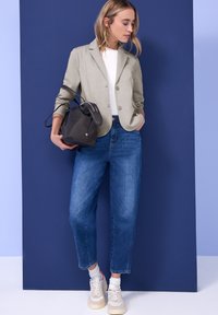 Femme en blazer beige, haut blanc, jean bleu et baskets blanches tenant un sac à main noir, debout devant un fond bleu.