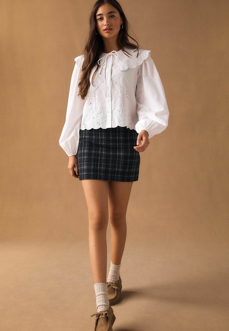 Bílá halenka s velkým volánovým límcem a průstřihy, v kombinaci s kari minisukní ve tmavých odstínech, podkolenkami po kolena a hnědými botami.