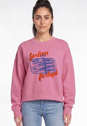 Femme portant un sweat-shirt rose avec un graphisme d'une boîte de sardines bleue et les mots "Sardines Portugal" en texte orange.