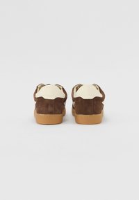 Baskets en suede marron avec des accents crème et une semelle en caoutchouc marron clair. Présentent des formes arrondies et un design minimaliste.